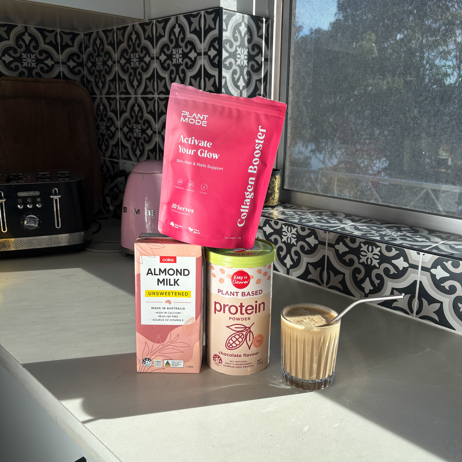 Supplement packages and a glass of a blended drink on a kitchen counter with a window in the background.